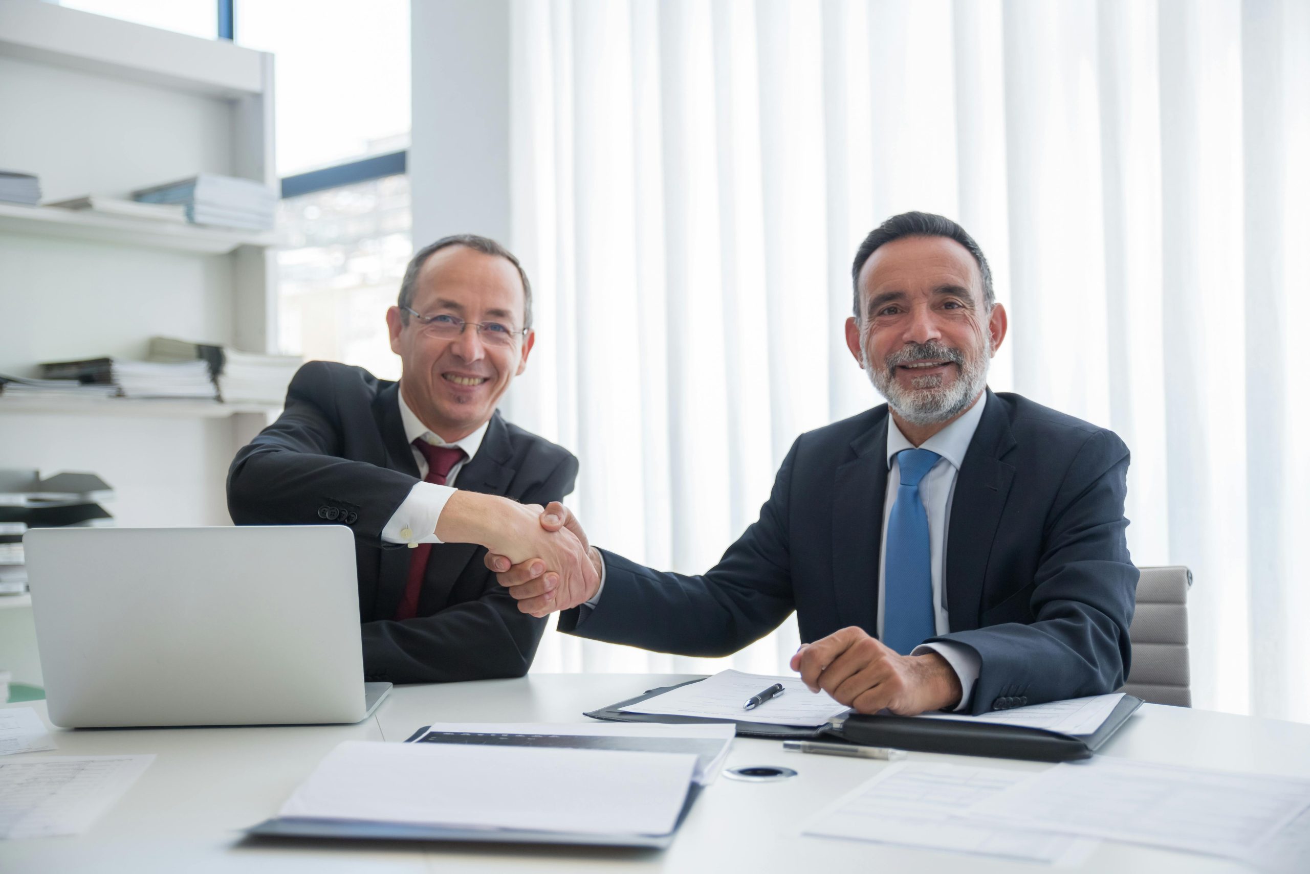 Two businessmen in formal suits shaking hands across a table in an office setting.